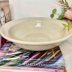 May include: A cream-colored, shallow ceramic bowl with a wide rim. The bowl is sitting on top of a stack of colorful books. A vase of pink tulips and greenery is to the right of the bowl. A book with Reese Witherspoon on the cover is to the left.