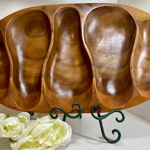 May include: A wooden serving tray with five oval compartments. The tray is made of a light brown wood with a glossy finish. It is displayed on a dark green metal stand, with white flowers in the foreground. This tray is perfect for serving snacks or appetizers.