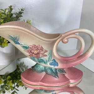 May include: A vintage ceramic gravy boat in a soft pink and cream color scheme. The boat features a floral design with pink and blue accents, and a decorative handle. The base is a matching pink color.