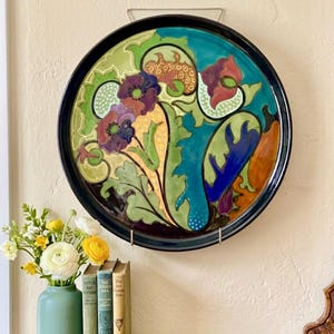May include: A large, round decorative plate with a black rim, featuring a floral design in shades of purple, red, green, and blue. The plate is mounted on a wall. A vase of flowers and books are on a wooden surface below.