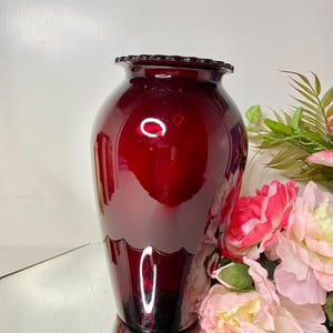 May include: A deep red glass vase with a smooth, rounded body and a scalloped rim. The vase features a reflective surface and a wide opening, showcasing a rich, dark crimson hue that catches the light, creating bright highlights.