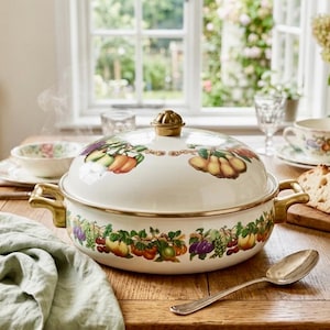 May include: A white enamel casserole dish with a lid, adorned with colorful fruit illustrations. The dish has gold-colored handles and trim. The table setting includes a spoon, bread, and a teacup.