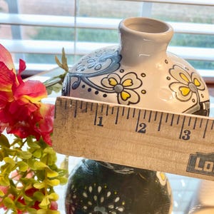 Puede incluir: Un peque&ntilde;o jarr&oacute;n decorativo de cer&aacute;mica con base blanca y dise&ntilde;o floral. El jarr&oacute;n mide aproximadamente 7,6 cm de di&aacute;metro. El dise&ntilde;o incluye detalles en azul, amarillo y negro. Una regla de madera se coloca delante del jarr&oacute;n.