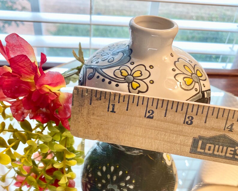 Puede incluir: Un peque&ntilde;o jarr&oacute;n decorativo de cer&aacute;mica con base blanca y dise&ntilde;o floral. El jarr&oacute;n mide aproximadamente 7,6 cm de di&aacute;metro. El dise&ntilde;o incluye detalles en azul, amarillo y negro. Una regla de madera se coloca delante del jarr&oacute;n.