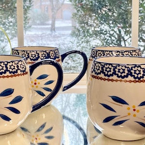 May include: Four ceramic mugs with a white base, adorned with blue floral patterns and dark blue handles. The mugs have a decorative border of blue and brown geometric shapes. The mugs are displayed on a mirrored surface.