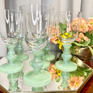 May include: A set of six clear wine glasses with light green stems and bases. The glasses are arranged on a mirrored tray, reflecting the light and the surrounding objects. A small gold-colored planter with flowers is in the background.