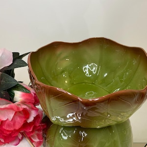May include: A ceramic bowl with a leaf design. The bowl has a glossy green interior and a brown rim. The exterior features a leaf pattern. The bowl is sitting on a mirrored surface, with pink and red flowers in the background.