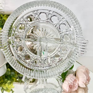 May include: A clear, divided glass serving dish with a starburst center design and geometric patterns. The dish has two handles and is elevated on a clear pedestal. The dish is likely used for serving snacks or appetizers.