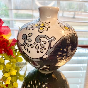 Puede incluir: Un peque&ntilde;o jarr&oacute;n redondo de cer&aacute;mica con cuello gris y un patr&oacute;n decorativo. El jarr&oacute;n est&aacute; dividido en dos secciones: una blanca con dise&ntilde;os florales grises y amarillos, y la otra negra con un patr&oacute;n de ondas.