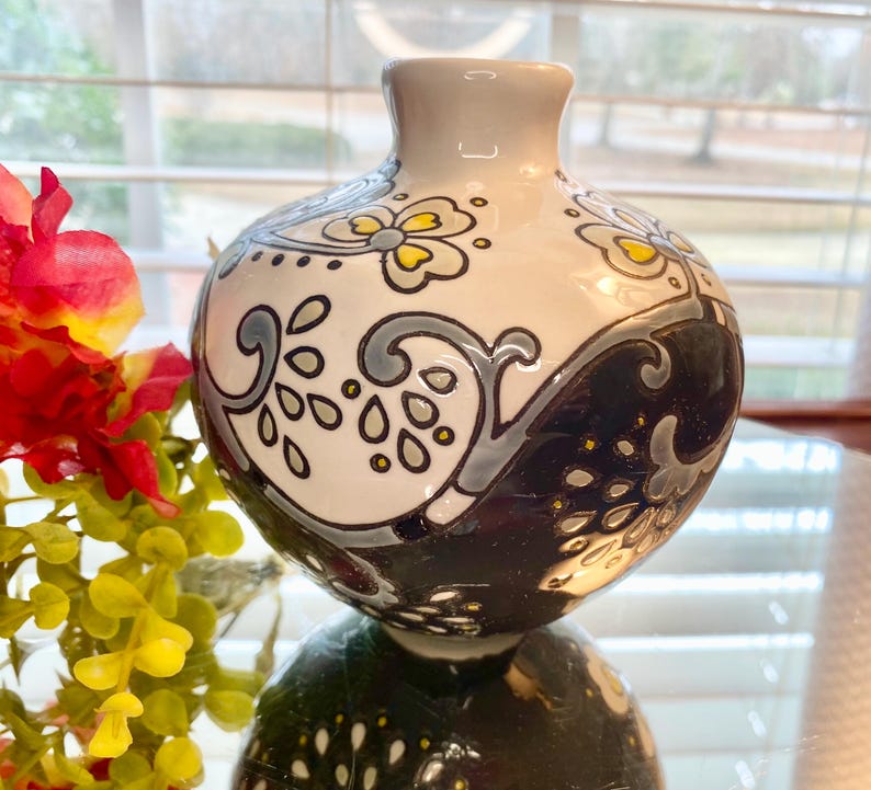 Puede incluir: Un peque&ntilde;o jarr&oacute;n redondo de cer&aacute;mica con cuello gris y un patr&oacute;n decorativo. El jarr&oacute;n est&aacute; dividido en dos secciones: una blanca con dise&ntilde;os florales grises y amarillos, y la otra negra con un patr&oacute;n de ondas.