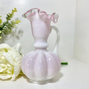 May include: A vintage pink glass pitcher with a ruffled rim and a white handle. The pitcher has a bulbous body with vertical ridges and a white base. The rim is trimmed with a delicate pink color.