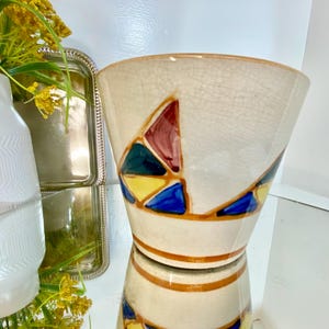 May include: Cream-colored ceramic vase with a geometric design featuring triangles in red, blue, yellow, and green. The vase has a wide opening and a brown rim. The vase is reflected in the surface below.