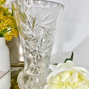 May include: A clear, cut-glass vase with a flared top and a round base. The vase features intricate starburst and geometric patterns. The vase is displayed with yellow flowers and a white flower in the background.