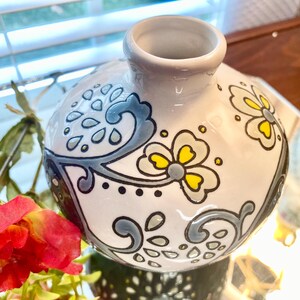 Puede incluir: Un jarr&oacute;n de cer&aacute;mica blanco con cuerpo redondo y cuello estrecho. El jarr&oacute;n est&aacute; decorado con motivos florales y abstractos grises y amarillos. El jarr&oacute;n est&aacute; sobre una superficie reflectante.