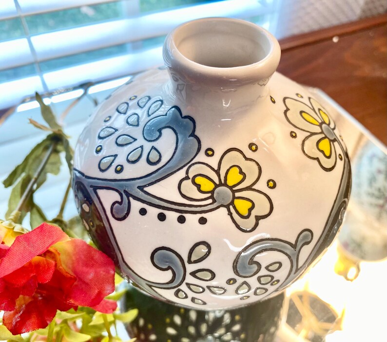 Puede incluir: Un jarr&oacute;n de cer&aacute;mica blanco con cuerpo redondo y cuello estrecho. El jarr&oacute;n est&aacute; decorado con motivos florales y abstractos grises y amarillos. El jarr&oacute;n est&aacute; sobre una superficie reflectante.