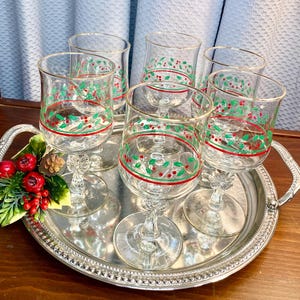 May include: A set of six clear wine glasses with a festive red and green holly berry design, arranged on a silver serving tray. The glasses have a decorative stem and are ready for a holiday celebration.
