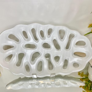 May include: An oval-shaped white ceramic tray featuring a series of irregular, organic-shaped cutouts throughout its surface. The tray has a glossy, smooth finish and a wavy, scalloped edge, creating a porous, open-work design across the entire piece.