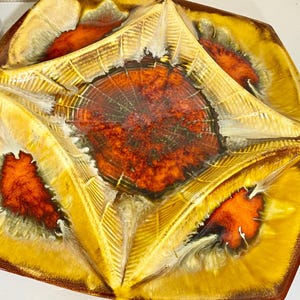 May include: A decorative ceramic serving dish with a unique design. The dish features a central reddish-brown area surrounded by yellow and orange sections. The dish has a textured surface and a handle on each side.