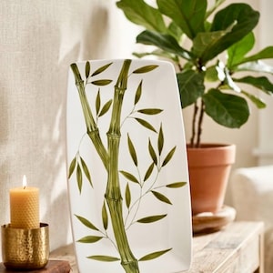 May include: A rectangular white ceramic serving platter featuring a hand-painted green bamboo stalk design with detailed leaves. The platter has slightly curved edges and a smooth, glossy finish, showcasing a botanical illustration.