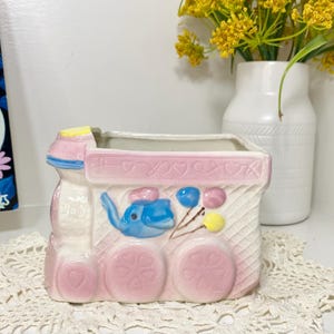 May include: A ceramic planter shaped like a train, primarily pink and white, with blue and yellow accents. The train features a blue elephant, pink wheels, and decorative balloons. The planter is designed to hold small plants or flowers.