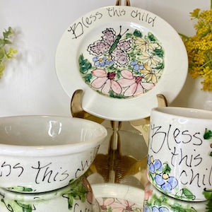 May include: A white ceramic plate, bowl, and mug set. The plate features a butterfly and floral design with the text "Bless this child." The bowl and mug also have the text "Bless this child" and floral accents.