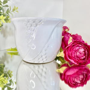 May include: A white ceramic planter with a tulip shape and a grape vine design. The planter is sitting on a reflective surface with pink flowers in the background.