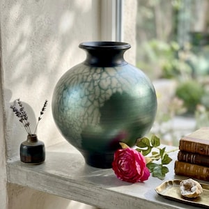 May include: A large, round ceramic vase with a wide body and a narrow neck, featuring a dark black rim and a gradient finish that transitions from deep forest green to a white, crackled, web-like texture across the surface of the rounded vessel.
