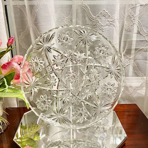 May include: A clear glass plate featuring a prominent starburst pattern with multiple etched star motifs radiating from the center. The circular plate has a scalloped edge and is displayed upright on a clear stand, showcasing intricate geometric cuts.