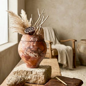 May include: A decorative, round, marbled vase in shades of brown and orange, filled with dried pampas grass, twigs, and a seed pod. The vase sits on a stone block on a wooden table, next to a leather-bound journal and a gold pen.