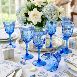 May include: A table setting with blue stemware, a floral centerpiece, and white linens. The blue glasses have a decorative pattern. A silver tray holds some of the glasses. White and blue flowers are in a vase.