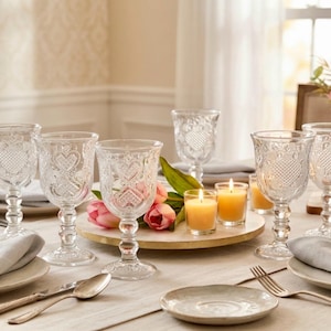 May include: A set of clear glass goblets featuring an embossed heart pattern and a textured stem. The goblets are arranged on a dining table alongside light gray cloth napkins, silver cutlery, small lit candles, and pink tulips in a vase.