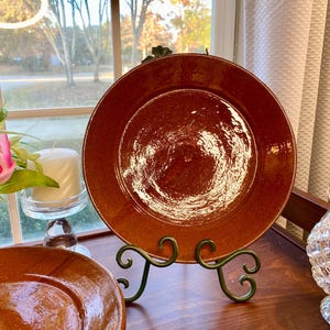 May include: A large, round, reddish-brown ceramic plate displayed on a decorative green metal stand. The plate has a glossy finish and is set against a window with a view of trees and a candle.
