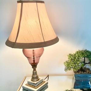 May include: A vintage table lamp with a beige lampshade and a pink glass base. The lamp has a brass stem and a marble base. A small bonsai tree in a square pot sits on the mirrored surface.