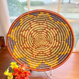 May include: A round, woven basket with a spiral design. The basket features concentric circles in shades of brown, yellow, and orange. The rim is a darker brown, and the basket is displayed on a clear stand.