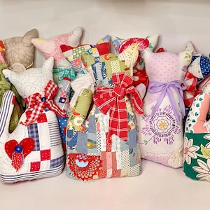 May include: A collection of handmade fabric cat-shaped pillows in various colors and patterns. Each pillow features unique patchwork designs, bows, and decorative elements, creating a charming and whimsical aesthetic. The pillows are displayed on a white shelf.