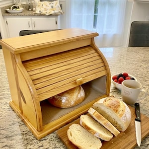 May include: A wooden bread box with a roll-top door, holding a loaf of bread. Sliced bread rests on a wooden cutting board next to the box. A mug and bowl of berries are also on the counter.