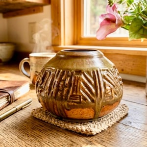 May include: A round, squat ceramic vase with a wide opening and a textured, relief-patterned surface. The pottery features a two-tone glaze with a dark, mossy green drip effect over a warm, speckled terracotta base, resting on a woven square mat.
