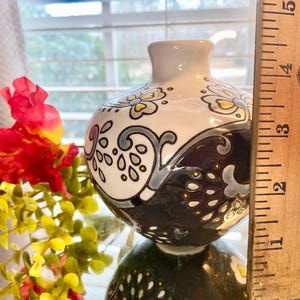 Puede incluir: Un peque&ntilde;o jarr&oacute;n decorativo de cer&aacute;mica con un dise&ntilde;o floral blanco, negro y gris. El jarr&oacute;n mide aproximadamente 10 cm de alto. El jarr&oacute;n tiene un cuerpo redondo y un cuello estrecho. El jarr&oacute;n est&aacute; junto a flores rojas y amarillas.