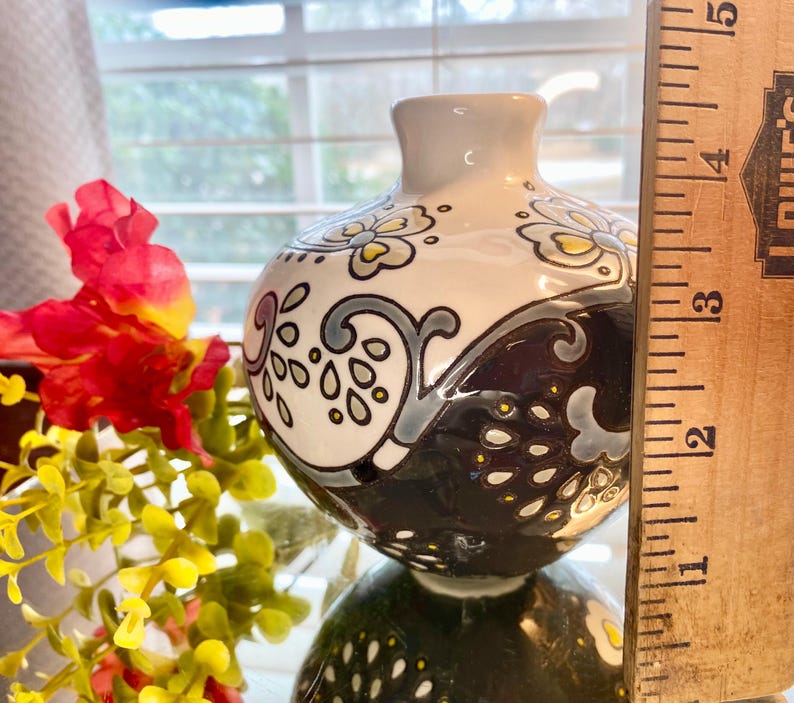 Puede incluir: Un peque&ntilde;o jarr&oacute;n decorativo de cer&aacute;mica con un dise&ntilde;o floral blanco, negro y gris. El jarr&oacute;n mide aproximadamente 10 cm de alto. El jarr&oacute;n tiene un cuerpo redondo y un cuello estrecho. El jarr&oacute;n est&aacute; junto a flores rojas y amarillas.