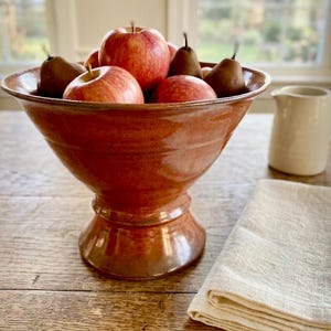 May include: A rustic, glazed terracotta fruit bowl filled with red apples and brown pears. The bowl sits on a wooden table, next to a folded cream-colored linen napkin and a small white pitcher. The bowl has a wide rim and a pedestal base.