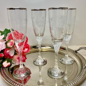 May include: Four clear champagne flutes with silver rims and etched floral designs. The stems have a twisted design, and the glasses are on a silver tray with a beaded edge. A bouquet of pink and red roses is in the background.