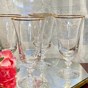 May include: A set of four clear crystal champagne flutes with gold rims. The glasses have a classic design with a flared bowl and a decorative stem. The flutes are arranged on a mirrored surface, with pink and red roses in the foreground.