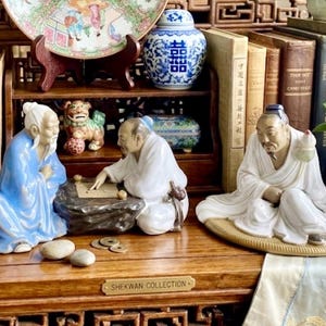 May include: Three porcelain figurines depicting figures in traditional robes. Two figures sit on a dark stone base playing a board game, while a third figure sits on a circular mat. The figurines are colored in white and blue, with a brass plaque reading SHEKWAN COLLECTION.