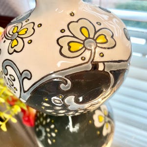 Puede incluir: Un jarr&oacute;n decorativo de cer&aacute;mica con acabado brillante. El jarr&oacute;n est&aacute; pintado con motivos florales en amarillo, gris y negro sobre un fondo crema. El jarr&oacute;n se refleja en una superficie espejada.