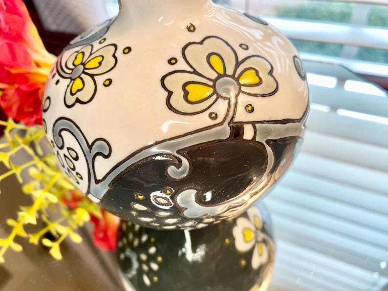 Puede incluir: Un jarr&oacute;n decorativo de cer&aacute;mica con acabado brillante. El jarr&oacute;n est&aacute; pintado con motivos florales en amarillo, gris y negro sobre un fondo crema. El jarr&oacute;n se refleja en una superficie espejada.
