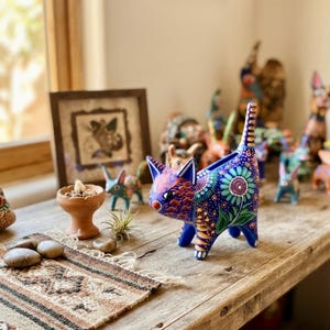 May include: A hand-painted ceramic cat figurine featuring a vibrant blue base color with intricate floral patterns in green, purple, and yellow. The cat has a long, upright tail, pointed ears, and detailed orange and blue markings on its face and body.