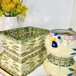 May include: A ceramic baking dish and a chicken-shaped pitcher. The baking dish is rectangular with a green floral pattern. The pitcher is cream-colored with blue, yellow, and green accents. A vase of yellow flowers is in the background.