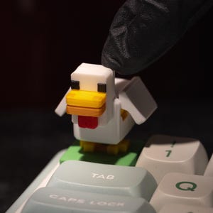 以下が含まれることがあります： 緑色の台座に乗った、白、黄色、赤のピクセル化されたニワトリの置物。パソコンのキーボードの上に置かれています。ニワトリは黄色いくちばしと赤い舌をしています。キーボードのキーは、白い文字が入った水色です。