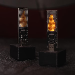 以下が含まれることがあります： 2つの電子的な炎の形をしたライト。各ライトは黒いベースとオレンジ色のLEDの長方形のディスプレイを備えています。LEDは炎のように配置されています。背景は暗いです。