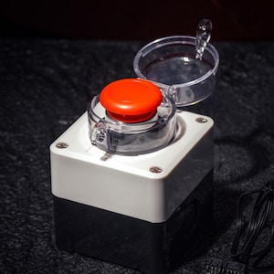 May include: A red button with a clear plastic cover sits on a white and black square base. The button is part of an electronic device, with a circuit board and black cable visible.
